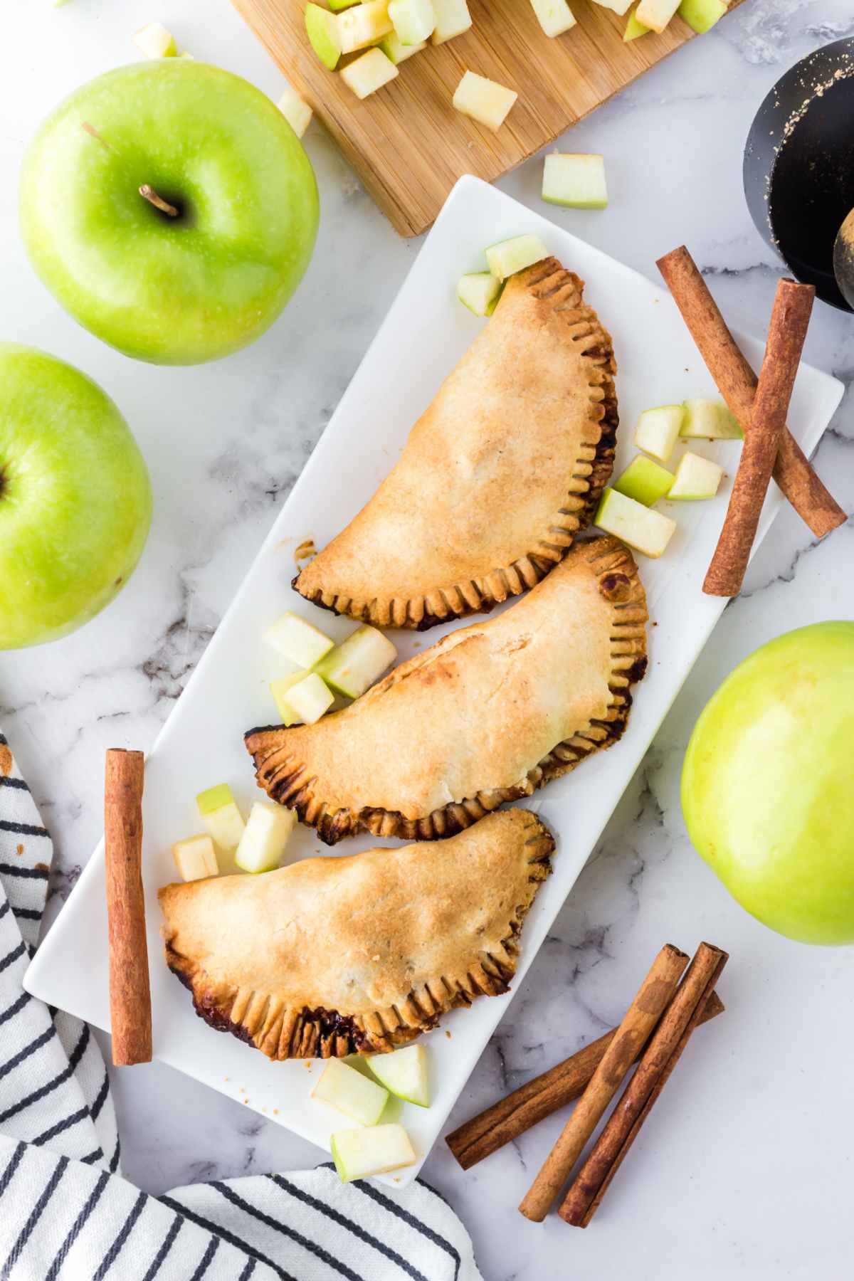 Three fully baked air fryer apple hand pies arranged on a white rectangular plate, surrounded by diced green apples and whole apples with cinnamon sticks scattered around.