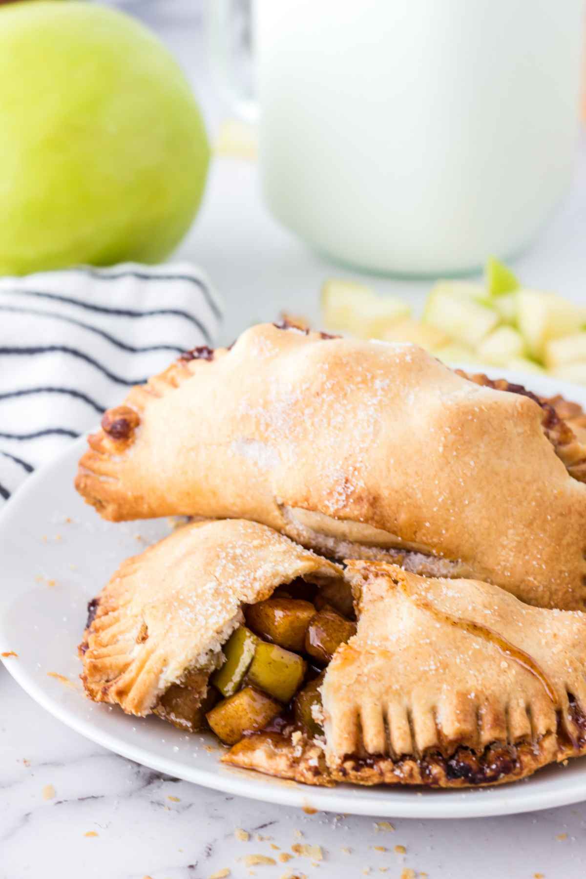 Two baked apple hand pies on a white plate, one cut open to reveal the cinnamon apple filling, with sugar crystals on top and a green apple blurred in the background.