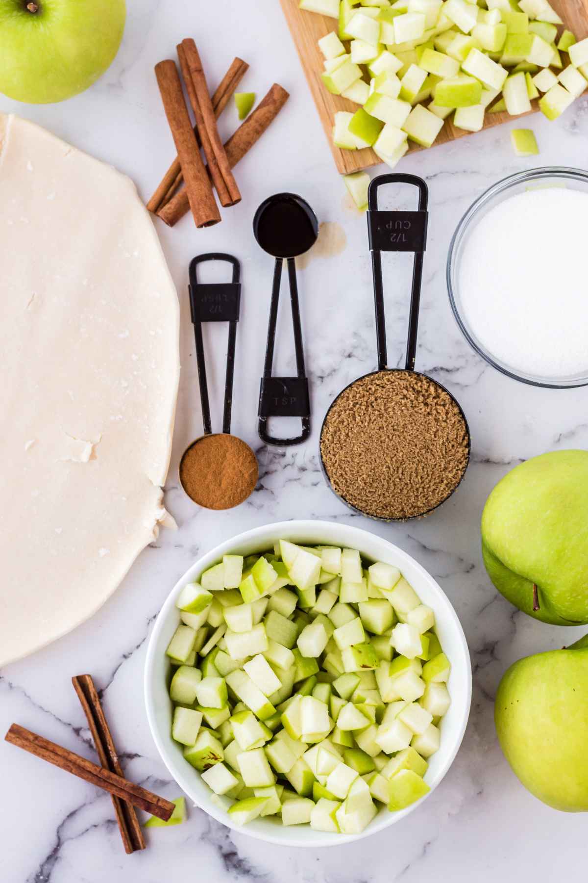 A flat lay of apple hand pie ingredients including diced green apples, pie crust dough, cinnamon sticks, brown sugar, white sugar, vanilla extract, and measuring spoons on a marble surface.