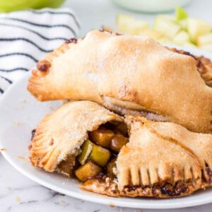 Two baked apple hand pies on a white plate, one cut open to reveal the cinnamon apple filling, with sugar crystals on top and a green apple blurred in the background.