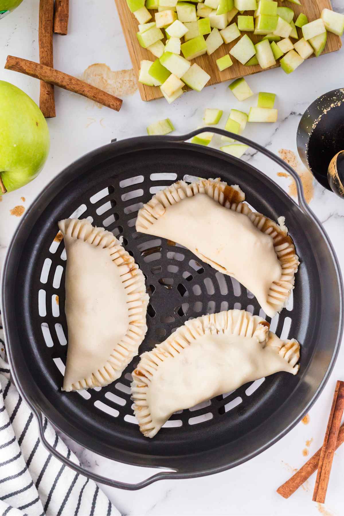 Three uncooked apple hand pies placed in an air fryer basket, edges sealed with fork marks, surrounded by diced apples, cinnamon sticks, and baking tools.
