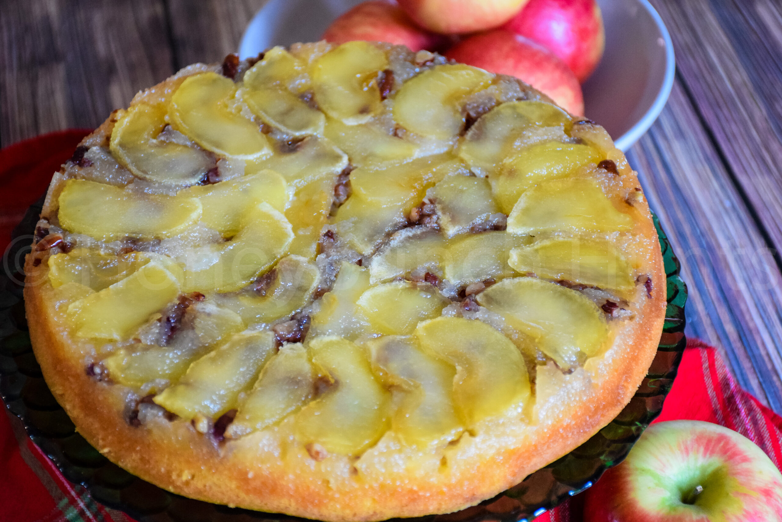 Angled close up of the apple upside down cake showing glossy apple slices and caramelized topping, with red apples in a bowl in the background.