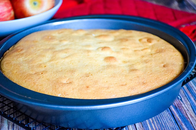 Baked white cake in a round pan with a lightly golden top, cooling on a wire rack over a wooden surface.