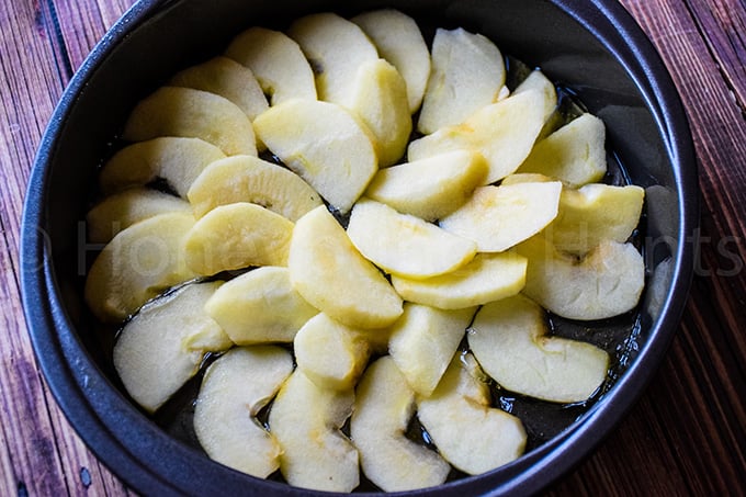 Sliced apples arranged in a circular pattern in a round cake pan, coated with a sugary glaze before baking.