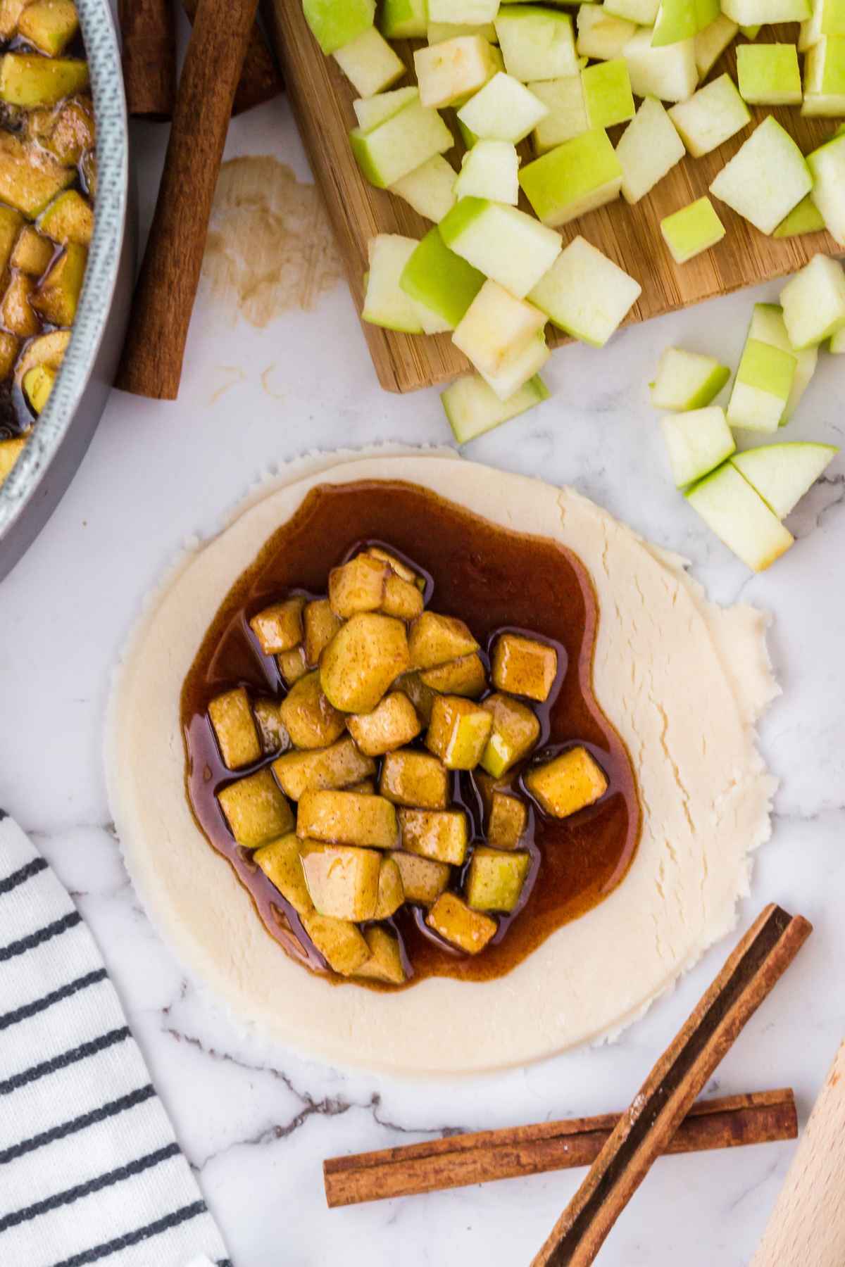 Close up of raw pie dough topped with a generous scoop of apple pie filling and thick cinnamon sauce, ready to be folded and sealed, surrounded by ingredients and cinnamon sticks.