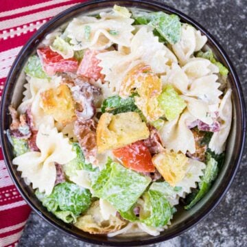 A bowl of BLT Pasta salad filled with bow tie pasta, chopped romaine lettuce, crispy bacon, grape tomatoes, red onion, and golden croutons, all tossed in a creamy dressing. A red and white striped towel is partially visible beside the bowl.