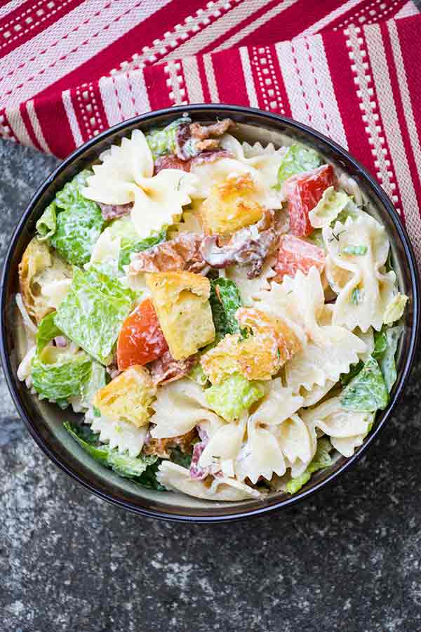 A bowl of BLT Pasta salad filled with bow tie pasta, chopped romaine lettuce, crispy bacon, grape tomatoes, red onion, and golden croutons, all tossed in a creamy dressing. A red and white striped towel is partially visible beside the bowl.