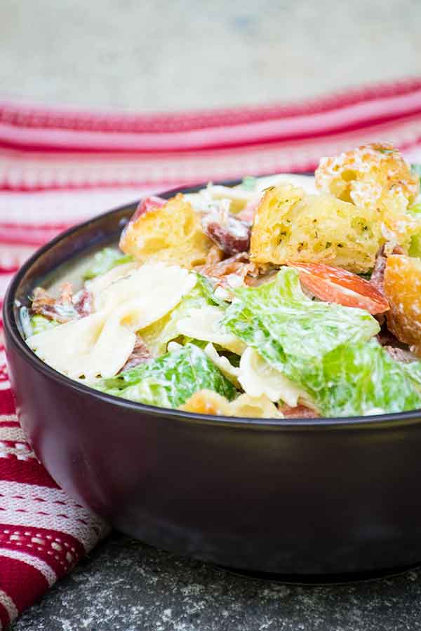 A close up side view of BLT Pasta salad in a dark bowl, showing crisp romaine lettuce, bow tie pasta, halved grape tomatoes, crumbled bacon, and golden homemade croutons, with a red striped cloth in the background.