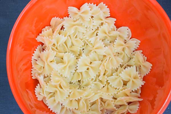 A bright orange bowl filled with cooked bow tie pasta, ready to be used as the base for a pasta salad recipe.