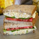 Close up of a stacked bacon egg salad sandwich on whole grain bread, layered with tomato slices and leafy greens.