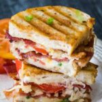 Stacked halves of a crispy grilled cheese sandwich filled with melted cheese, bacon, and tomato slices, garnished with chopped green onions on a white plate with a rustic edge. A few slices of fresh tomato and a bacon piece are on the side.