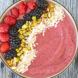 A thick, pink smoothie bowl topped with fresh raspberries, blackberries, chopped pistachios, and shredded coconut, arranged in diagonal rows.