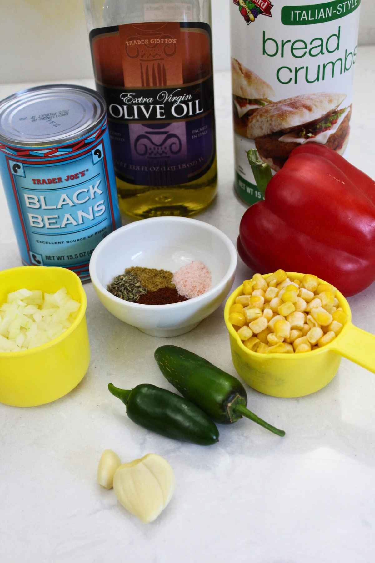 Ingredients displayed on a countertop, including canned black beans, olive oil, bread crumbs, red bell pepper, frozen corn, diced onion, jalapenos, garlic cloves, and assorted spices in a small white bowl.