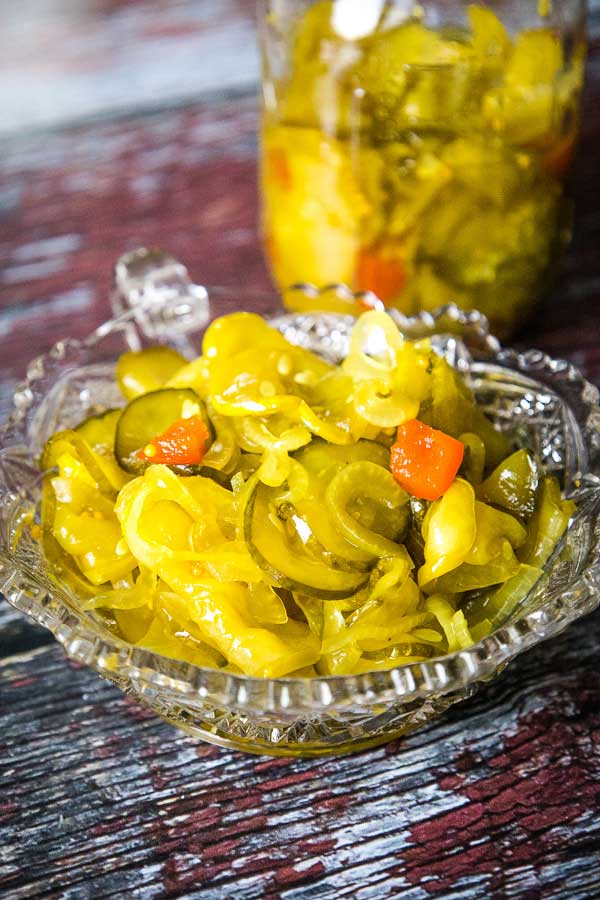 A clear glass dish with bread and butter pickles, including cucumber slices, onions, and red pepper chunks, with a jar in the background.