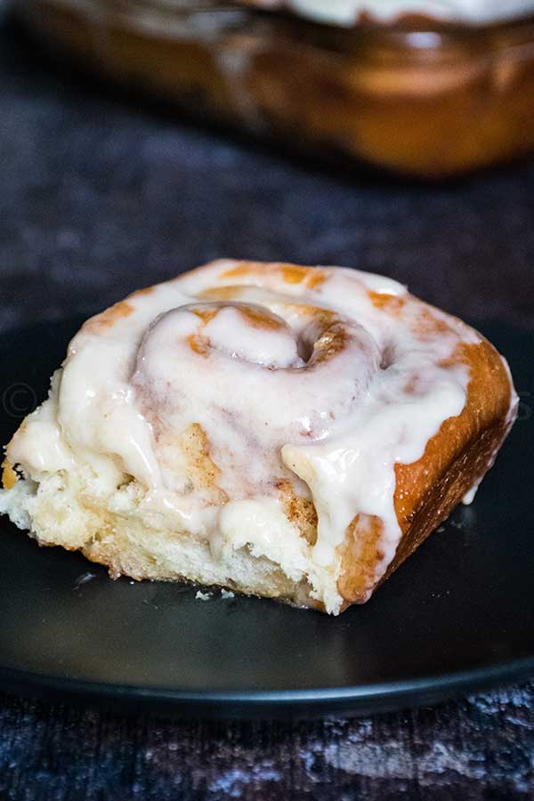 Single cinnamon roll on a dark plate, covered in icing with visible swirls and a soft fluffy interior.