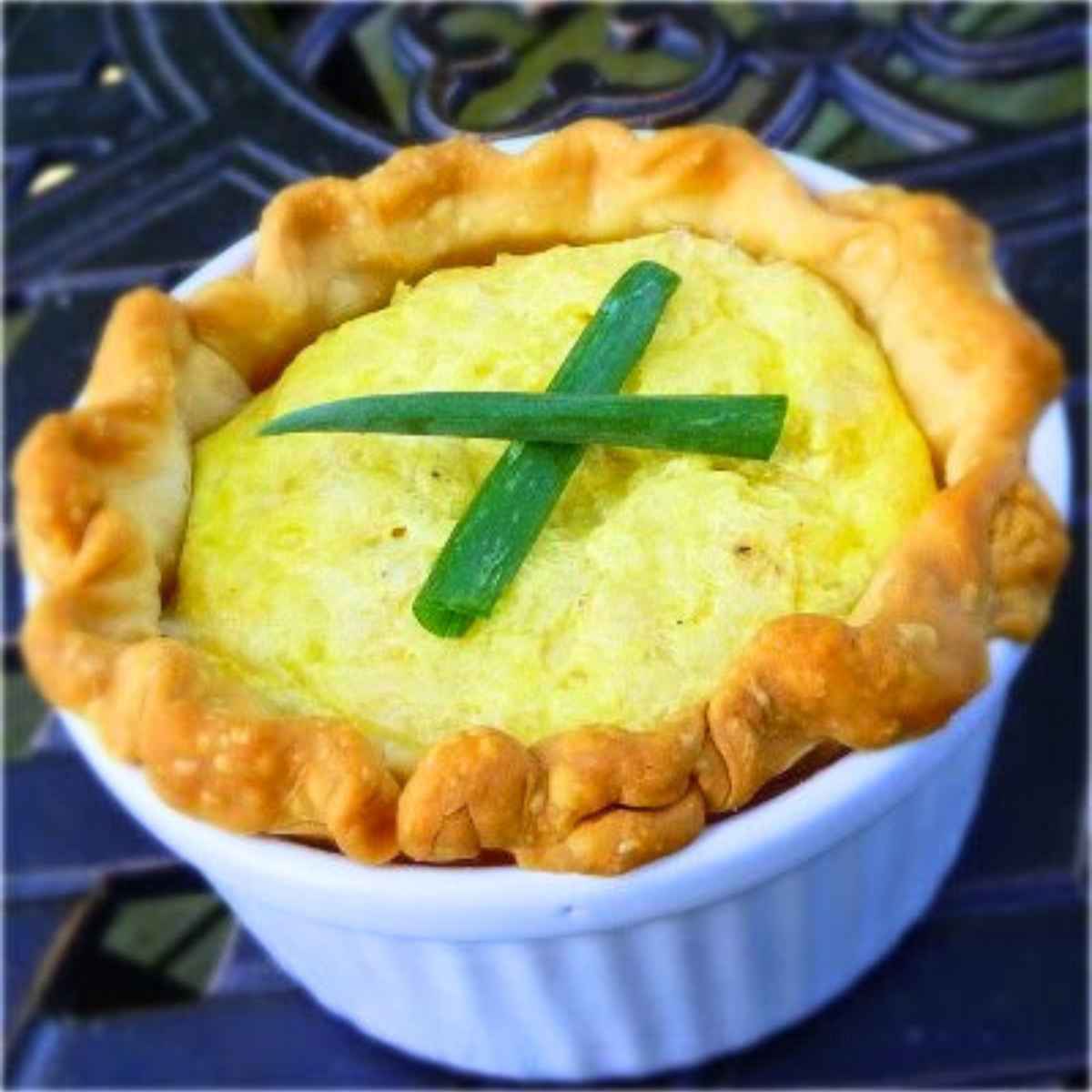 Individual Crab and Swiss Quiche in a white ramekin, topped with two green onion pieces in a crisscross pattern and surrounded by a golden, crimped pie crust.
