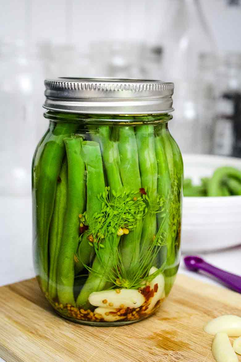 Side view of a glass jar packed with green beans, garlic cloves, dill, mustard seeds and red pepper flakes, sealed and placed on a wooden cutting board.