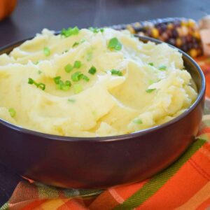 Creamy mashed potatoes garnished with chopped chives in a black bowl, set on a plaid autumn napkin.