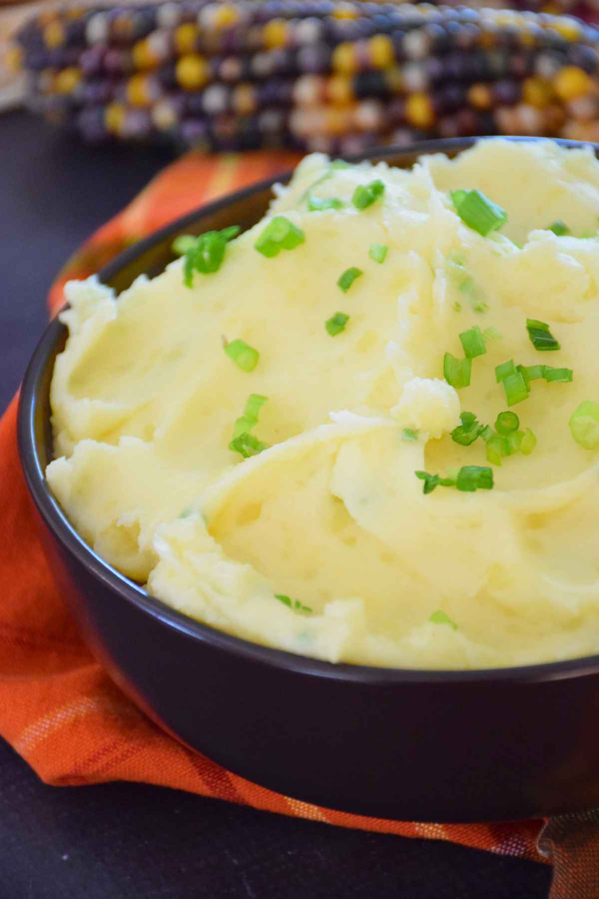 Close up overhead angled view of a bowl of mashed potatoes topped with chopped green chives, with a decorative ear of corn in the background.