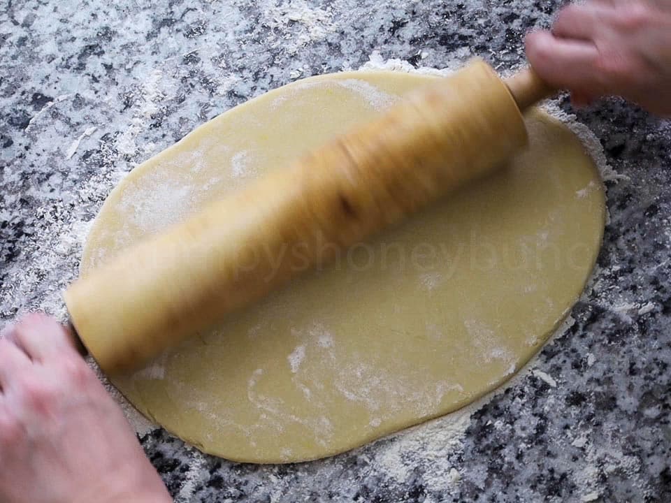 Rolling pin flattening cookie dough on a floured granite surface, hand visible in motion above.