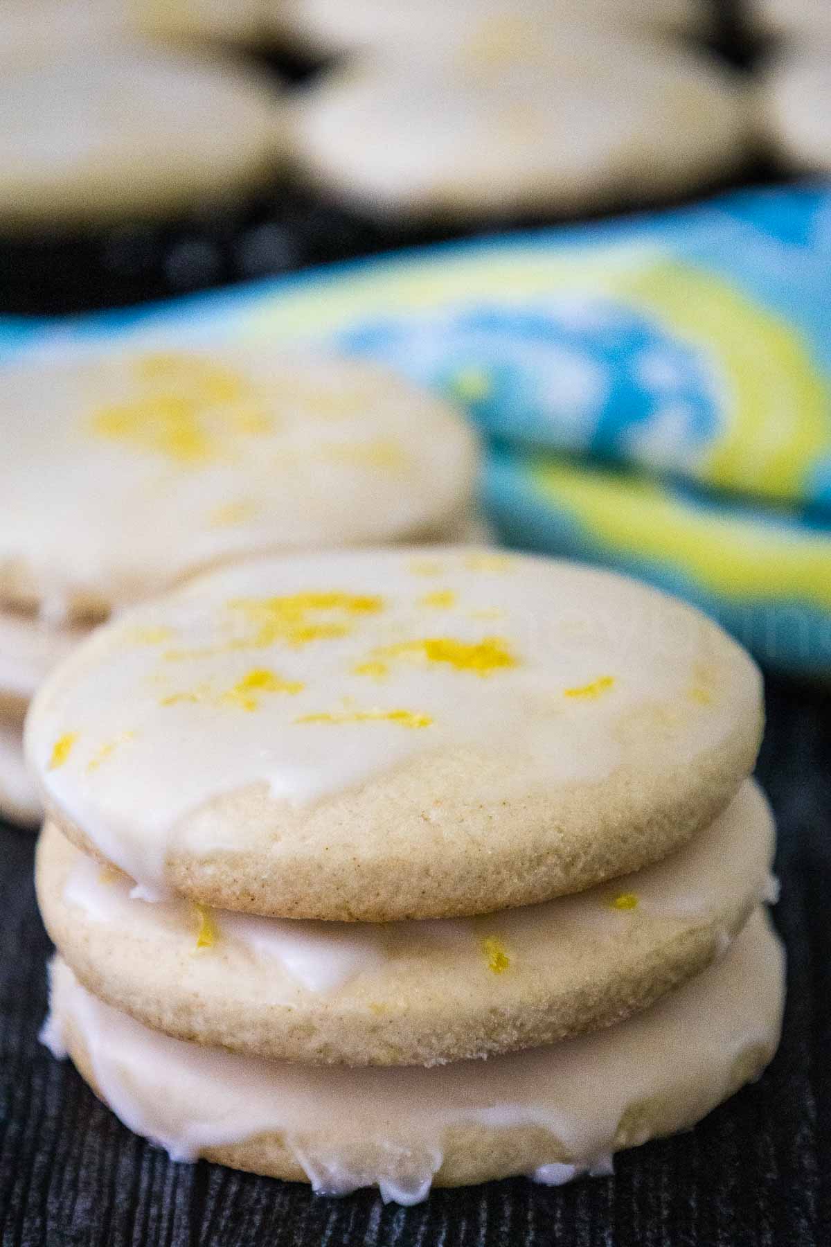 Stack of three glazed lemon cookies with a light icing and lemon zest on top, set on a dark wooden surface with more cookies in the background.