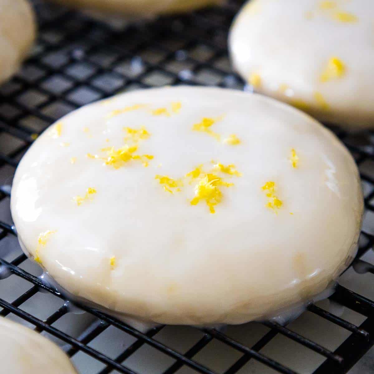 Glazed lemon cookie on a cooling rack with lemon zest sprinkled on top, surrounded by more cookies in soft focus.