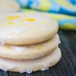 Stack of three glazed lemon cookies in sharp focus with colorful blue and yellow cloth blurred in the background.
