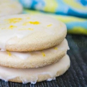 Stack of three glazed lemon cookies in sharp focus with colorful blue and yellow cloth blurred in the background.