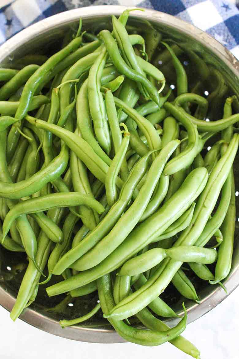 Colander filled with freshly washed green beans, sitting on a white countertop next to a blue and white checkered cloth.