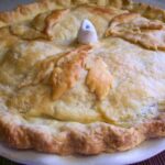 Whole baked apple pie with golden flaky crust, decorated with pastry leaf cutouts and a white ceramic pie bird vent in the center, sitting on a white pie dish.