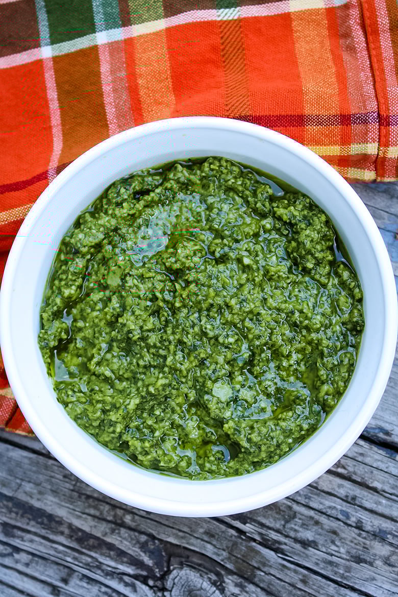 Overhead view of a white bowl filled with vibrant green homemade basil pesto, set on a rustic wooden surface with an orange and plaid cloth in the background.