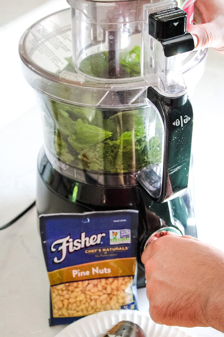 A food processor filled with fresh basil leaves being operated by hand, with a bag of Fisher brand pine nuts and a white plate in the foreground.