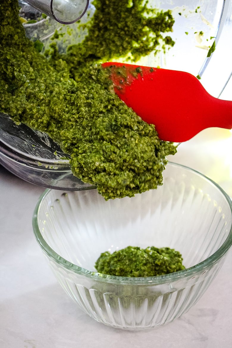 Homemade basil pesto being scraped from a food processor into a clear glass bowl using a red silicone spatula.
