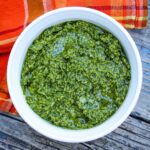 Overhead view of a white bowl filled with vibrant green homemade basil pesto, set on a rustic wooden surface with an orange and plaid cloth in the background.