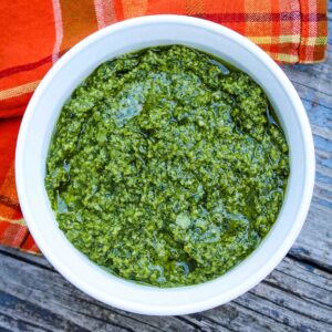 Overhead view of a white bowl filled with vibrant green homemade basil pesto, set on a rustic wooden surface with an orange and plaid cloth in the background.