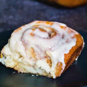 Close up of a frosted cinnamon roll on a dark plate, showing its tender crumb and gooey center.
