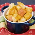 A small navy blue bowl with handles filled with golden, crispy homemade croutons, placed on a colorful red and green cloth.