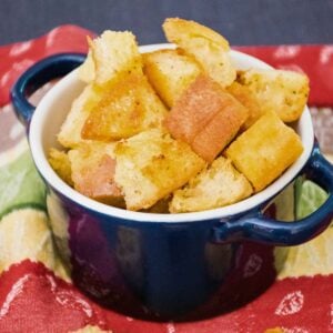 A small navy blue bowl with handles filled with golden, crispy homemade croutons, placed on a colorful red and green cloth.