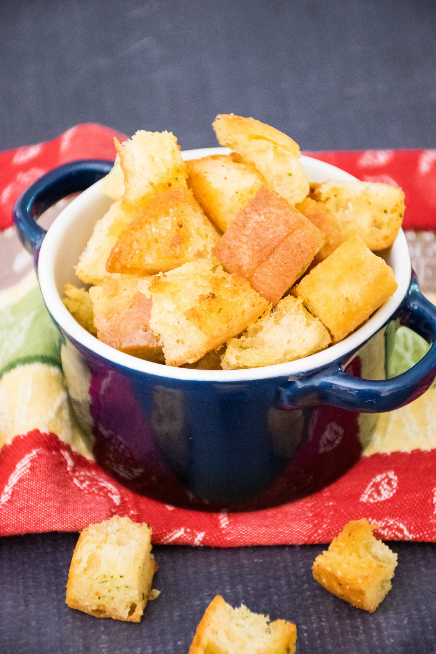 Am navy blue bowl filled with golden, crispy, homemade croutons sits on a colorful red, green, and cream striped kitchen towel with a few croutons scattered on the dark countertop around the bowl.