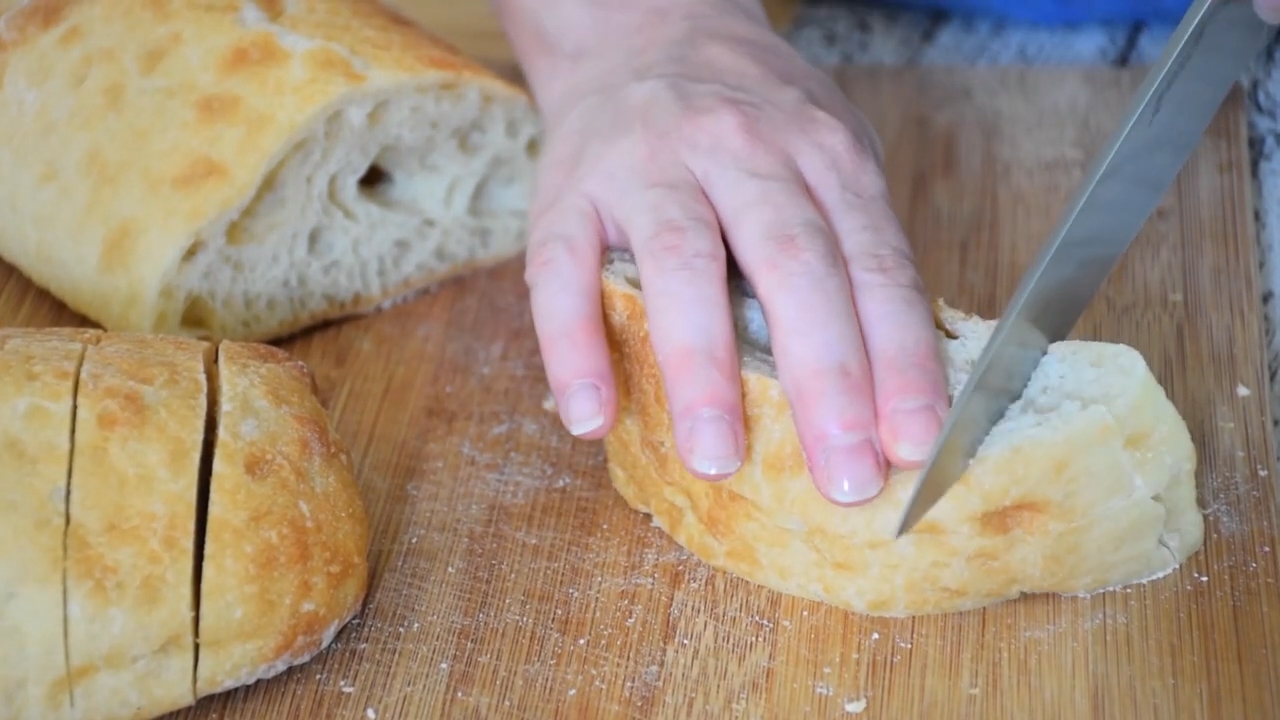 A hand holds a loaf of ciabatta bread steady on a wooden cutting board while slicing it with a serrated knife, preparing the bread for homemade croutons.