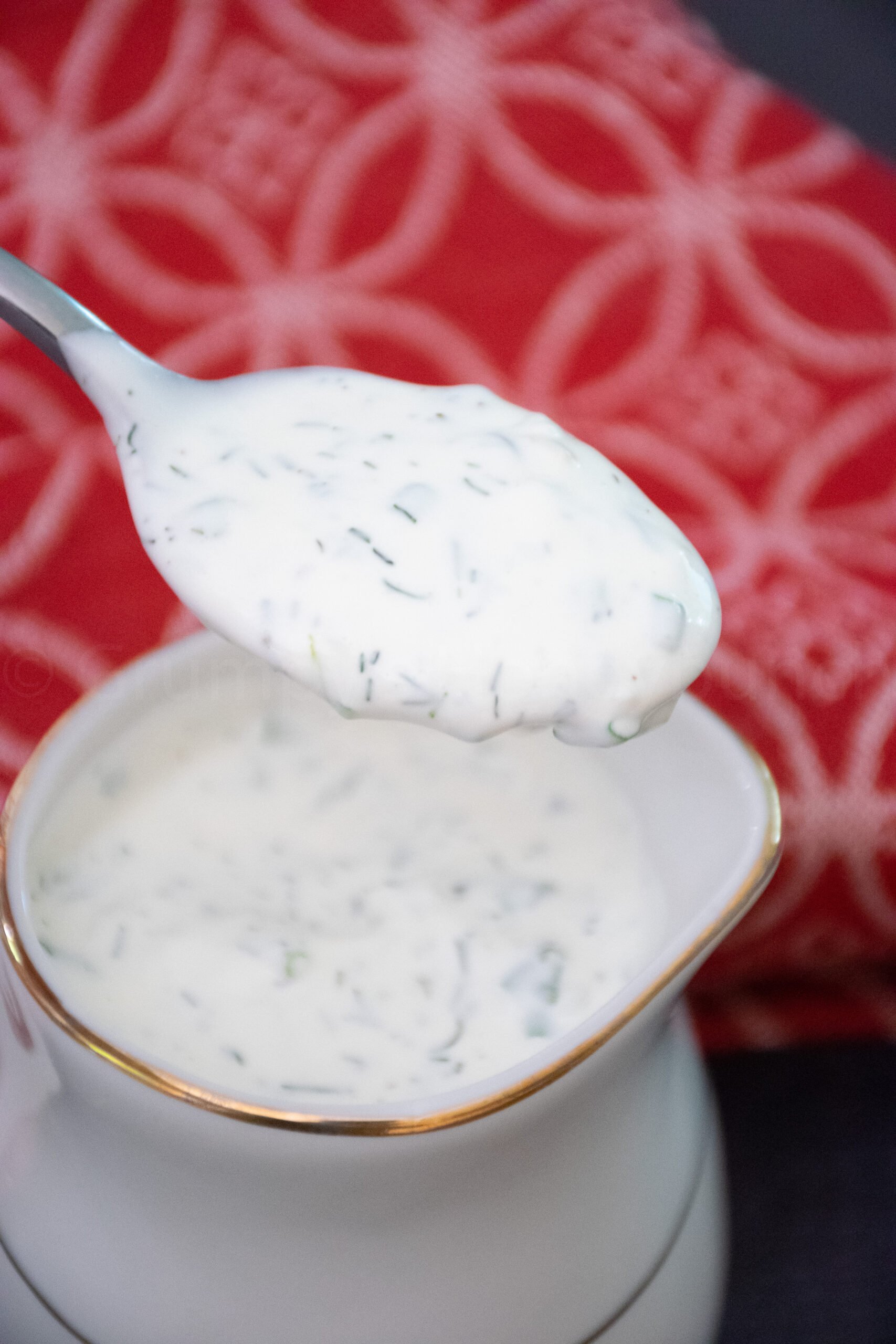 A spoonful of creamy homemaderanch dressing, speckled with fresh herbs, is held above a white and gold rimmed sauce dish filled with more dressing. A red patterned cloth is in the background.