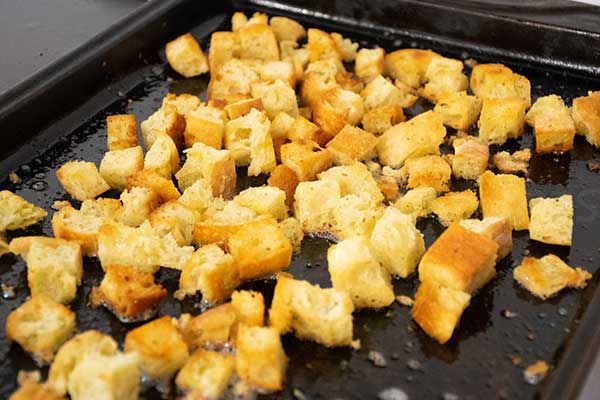 Browned cubes of buttery cubes of bread spread out on a dark baking sheet, golden and crisp from roasting with a light sheen of oil visible on the pan.