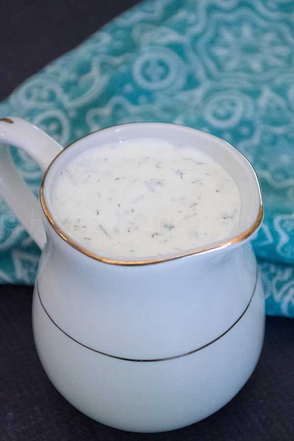 A small white pitcher with gold trim filled with homemade ranch dressing, speckled with herbs. A teal patterned cloth is softly draped in the background.