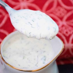 A spoonful of creamy homemaderanch dressing, speckled with fresh herbs, is held above a white and gold rimmed sauce dish filled with more dressing. A red patterned cloth is in the background.