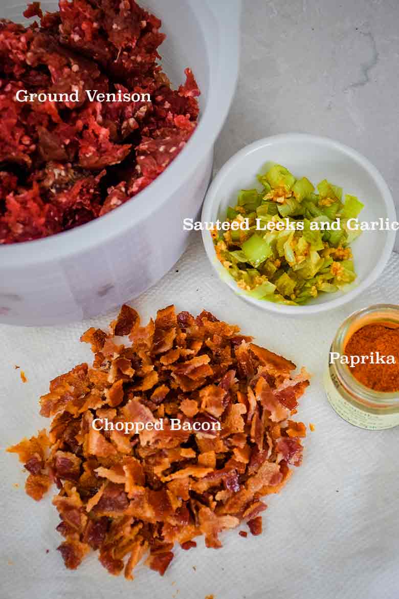 Overhead image of labeled ingredients for venison meatballs, including a bowl of ground venison, a small dish of sauteed leeks and garlic, a pile of chopped cooked bacon, and a jar of paprika.