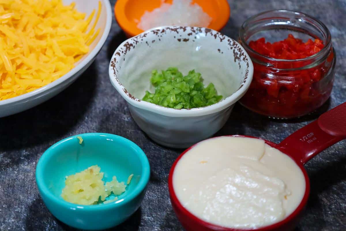 Assorted ingredients in a variety of bowls arranged on a dark countertop, including a bowl of shredded cheddar cheese, a small bowl of chopped green jalapenos, a jar of diced pimentos, a measuring cup filled with mayonnaise, a small dish of minced garlic, and another bowl containing chopped onions.