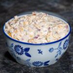 A bowl of creamy jalapeno pimento cheese spread, filled with shredded cheddar, diced pimentos, and bits of green jalapeno, served in a white ceramic bowl with blue floral designs on a dark countertop.