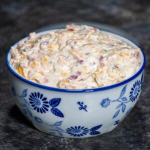A bowl of creamy jalapeno pimento cheese spread, filled with shredded cheddar, diced pimentos, and bits of green jalapeno, served in a white ceramic bowl with blue floral designs on a dark countertop.