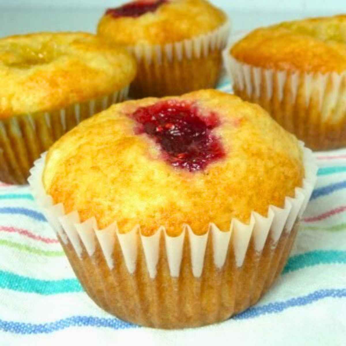 A group of golden brown muffins withjam filled centers. One muffin prominently showing a red jam resting on a colorful striped cloth.