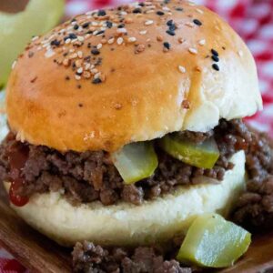 Loose meat sandwich served on a homemade bun topped with everything seasoning, filled with ground beef, dill pickles, and ketchup, with veggie fries in the background.